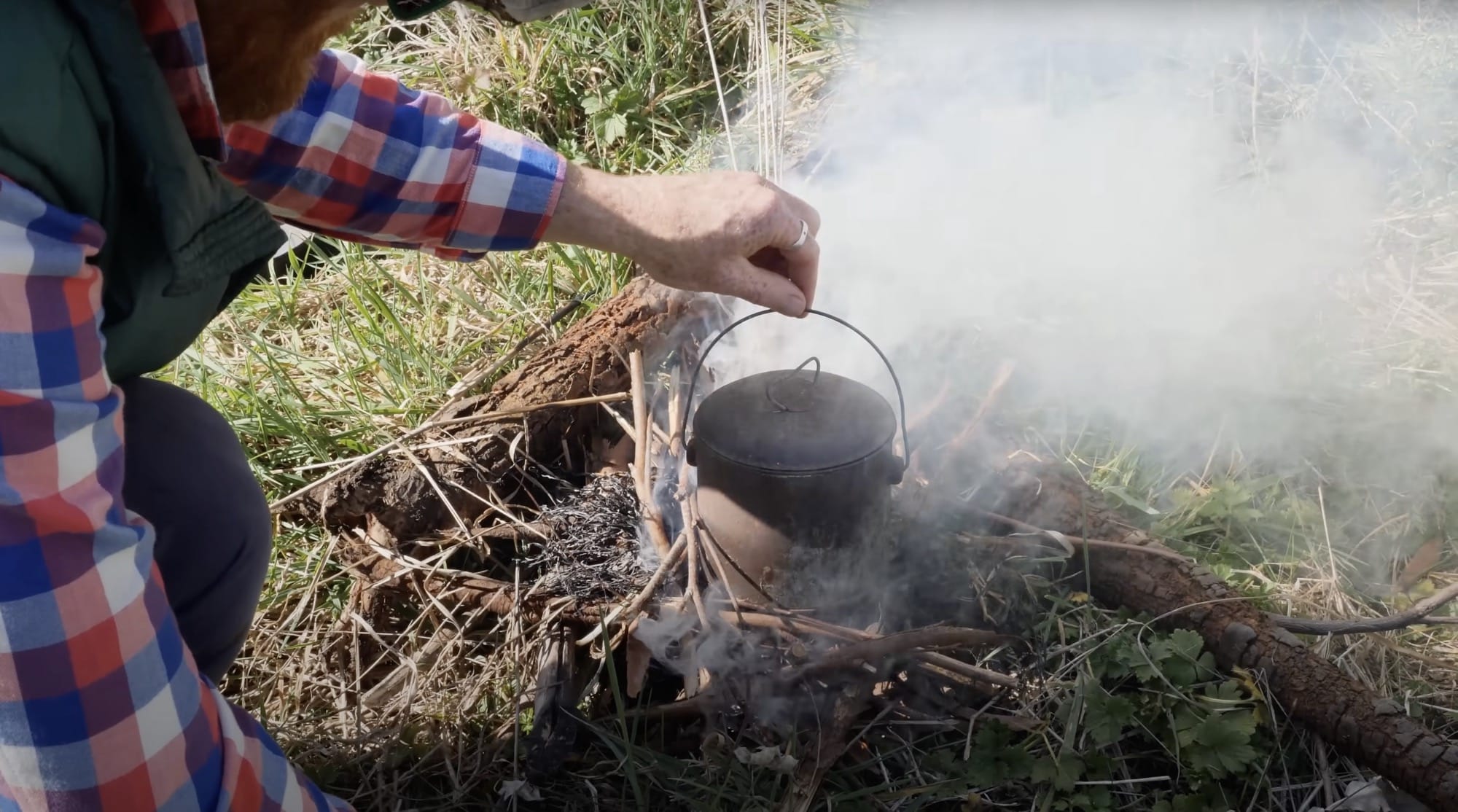 a still from a video showing a man's hand reaching over a small pot in a fire